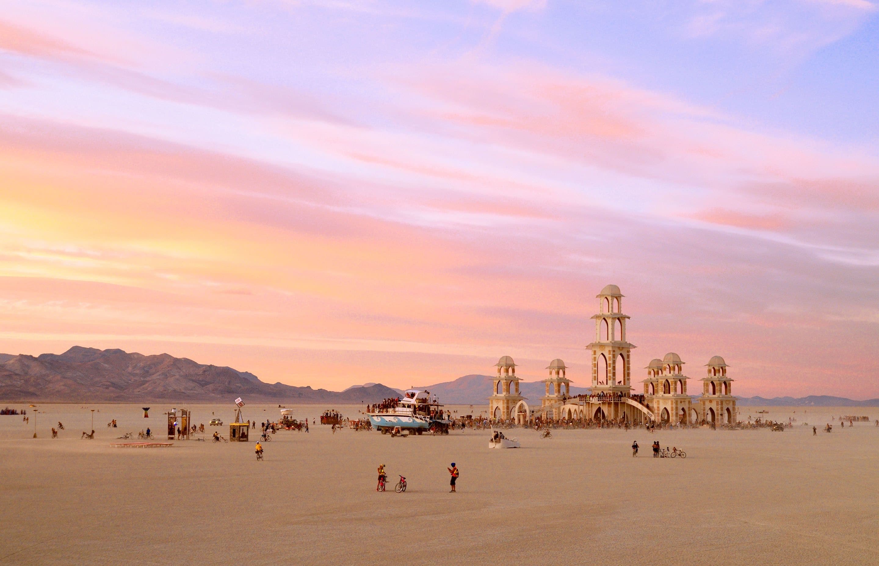 Three small figures with bikes far out on the open playa at pastel dusk, the temple on the horizon.