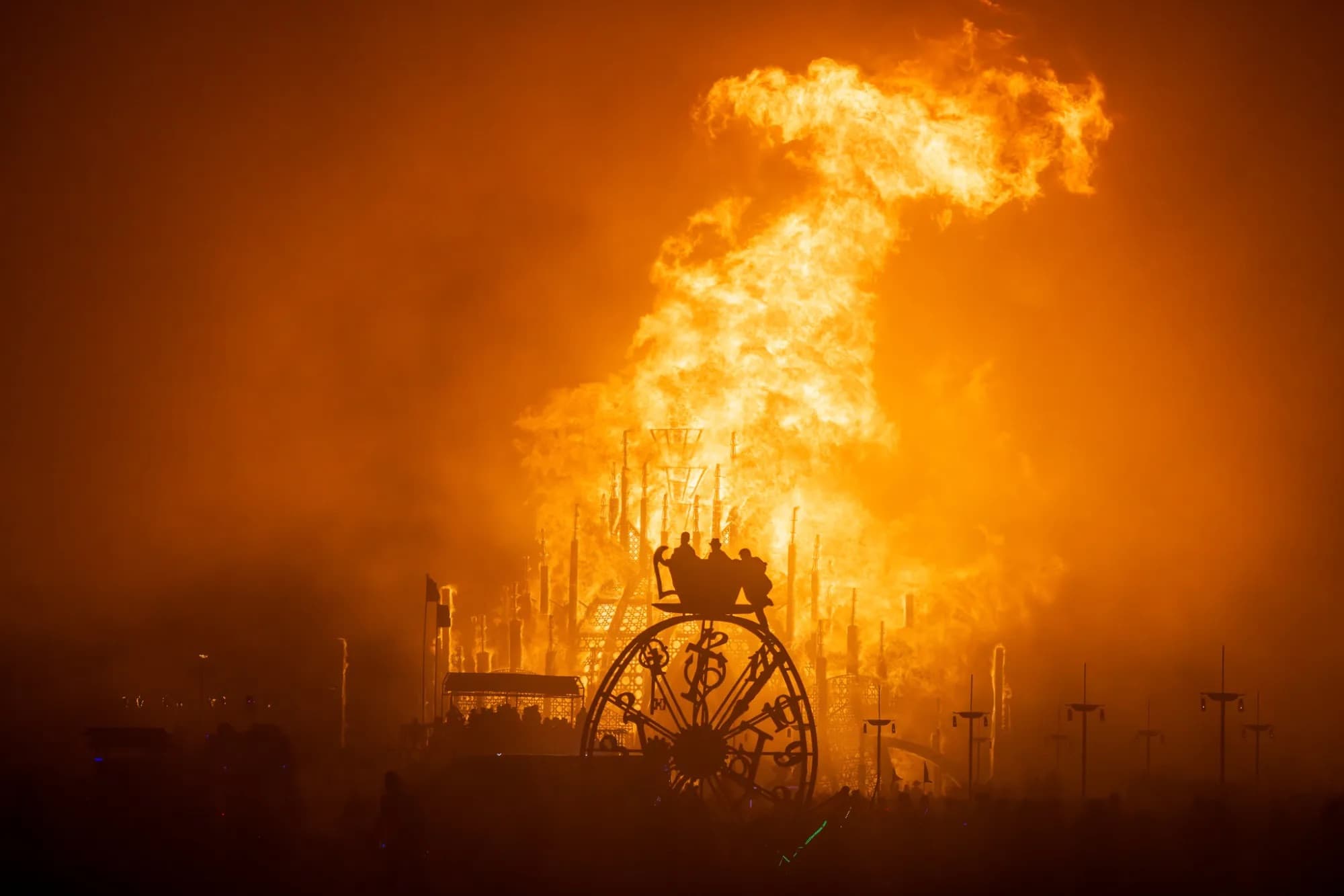 An orange column of fire rising behind the silhouette of a wheel structure on the open playa at night.