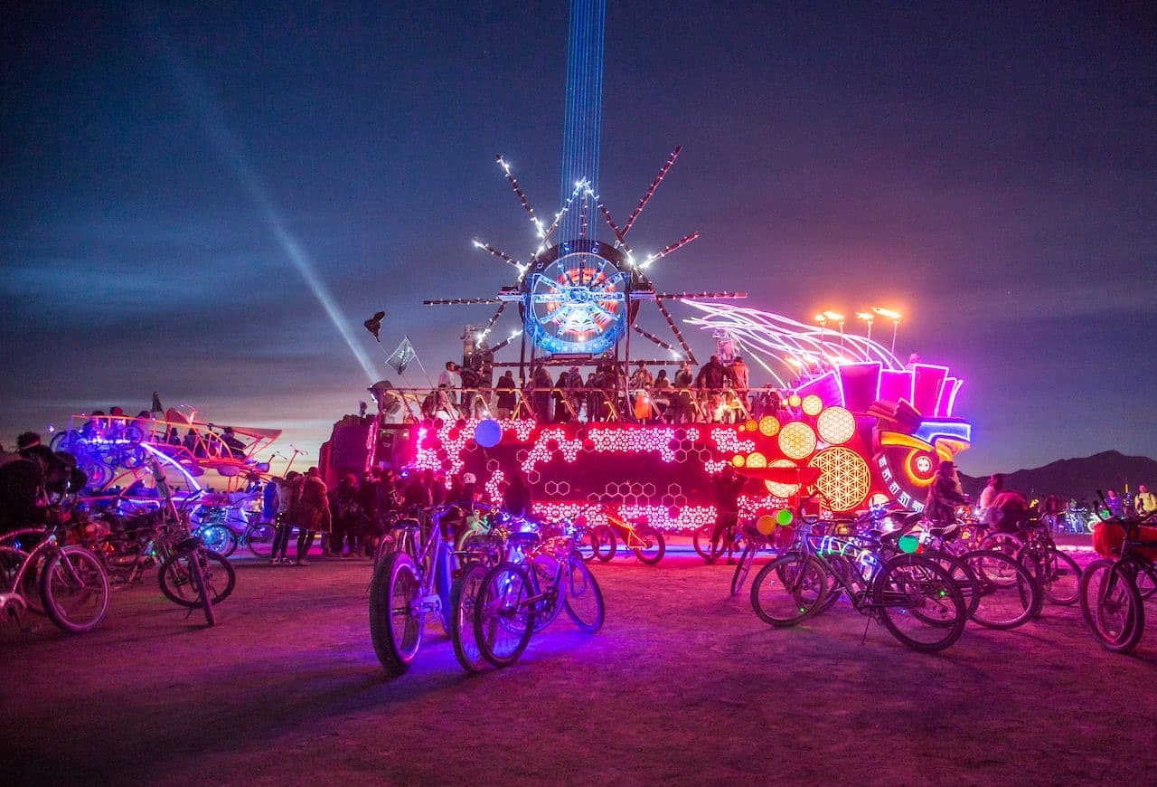A row of bikes with lit wheel rings parked in front of a neon art car at night on the playa.