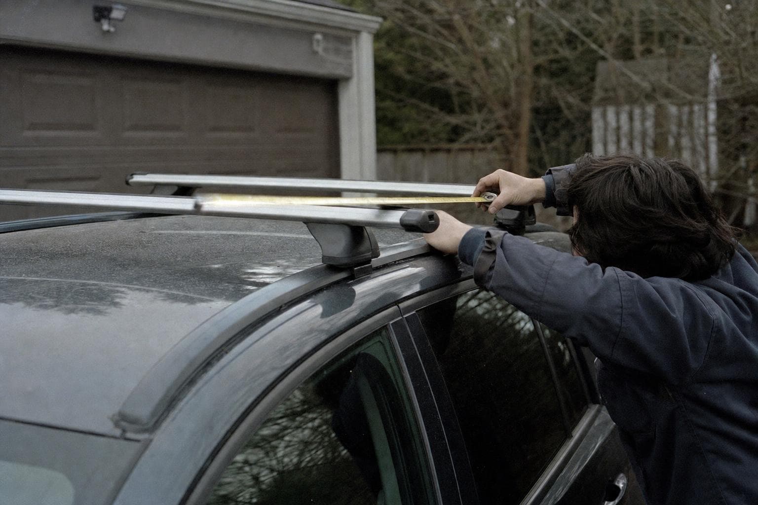 Hands running a tape measure along a roof crossbar in a garage, the car half loaded for the drive out.