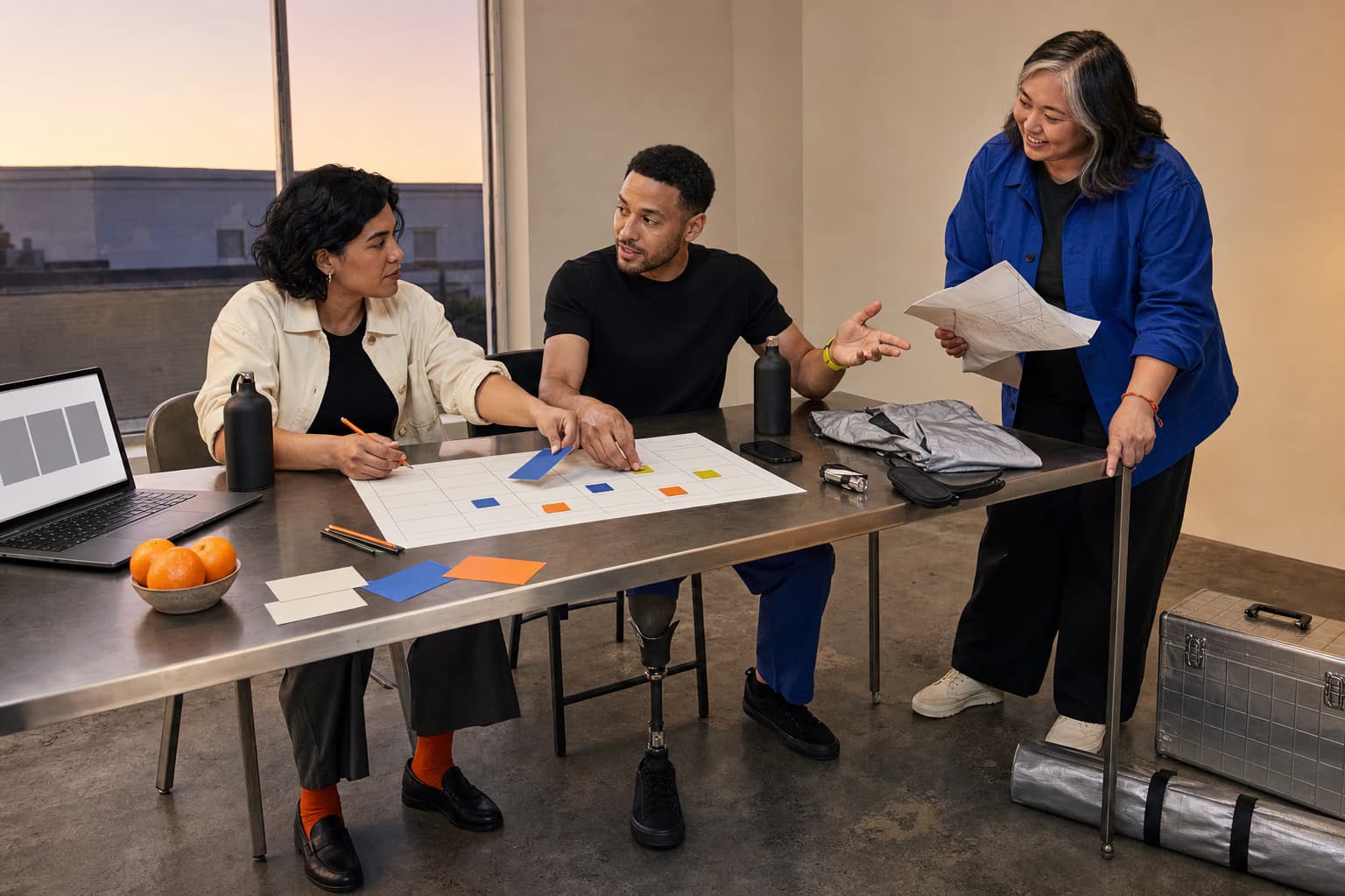 Three friends building a shared trip plan with a paper grid, gear tokens, textiles, and notes around a studio table
